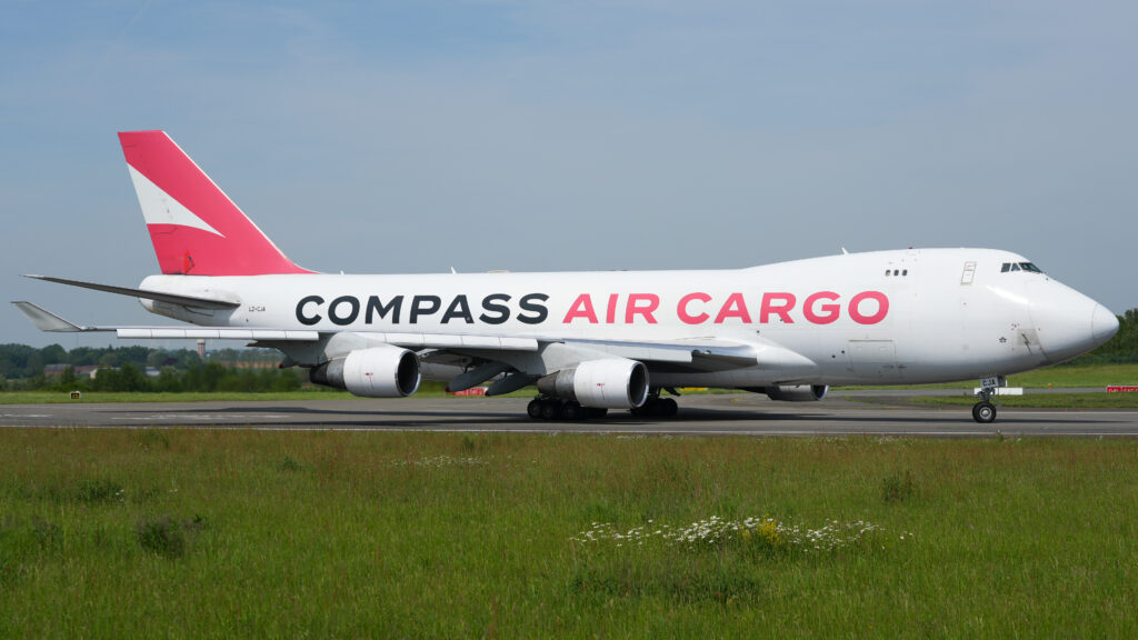 Boeing 747 on a runway. The aircraft is white with red and black text, set against a clear blue sky and grassy field.
