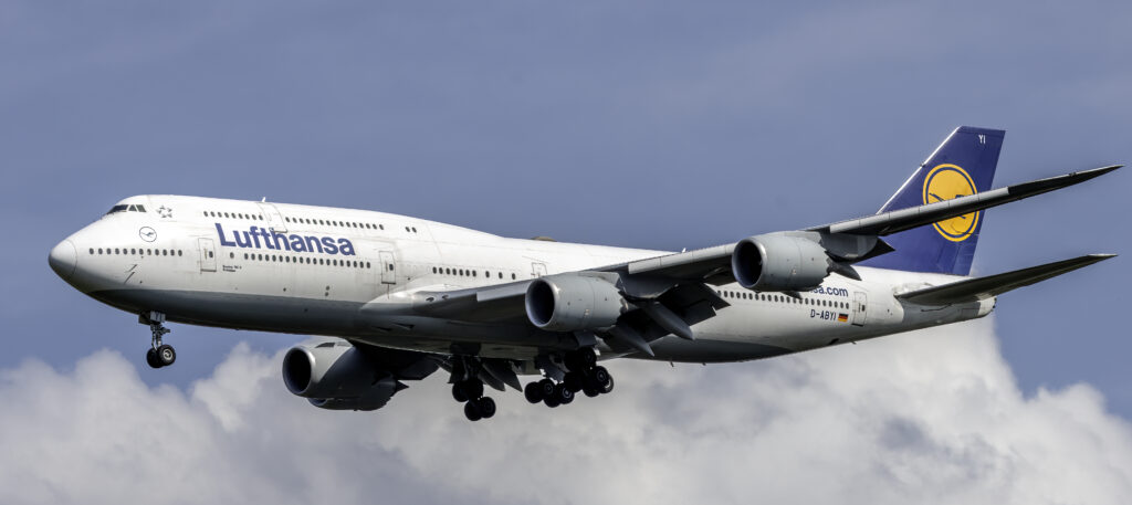 A Lufthansa Boeing 747 aircraft in flight against a backdrop of blue sky and clouds. The jet is descending, with landing gear visible.
