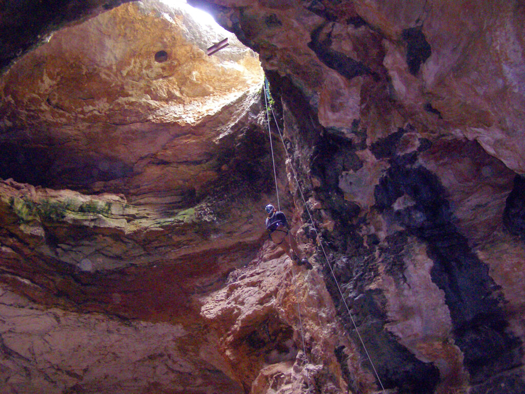 A climber is descending into a dimly lit cave, surrounded by rugged rock formations. Sunlight peeks through the cave opening, creating a dramatic scene.