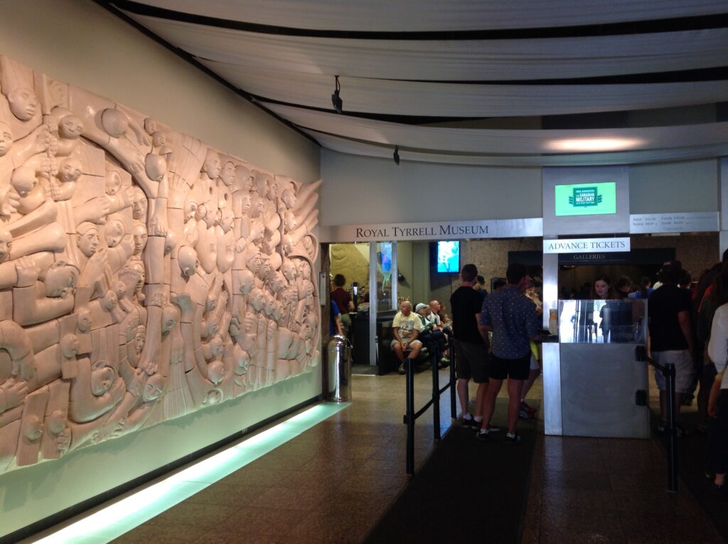 Queue at Royal Tyrrell Museum entrance with intricate wall sculpture. People wait for tickets, some seated, in a lively and curious atmosphere.