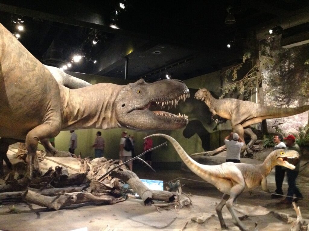 A museum exhibit displays lifelike dinosaur models, including a large Tyrannosaurus rex with mouth open and two smaller dinosaurs nearby. Visitors observe in the background.