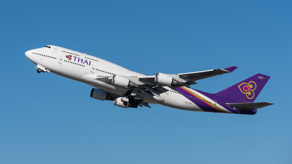 A Thai Airways Boeing 747 with a white body and purple tail ascends against a clear blue sky, conveying a sense of freedom and travel.