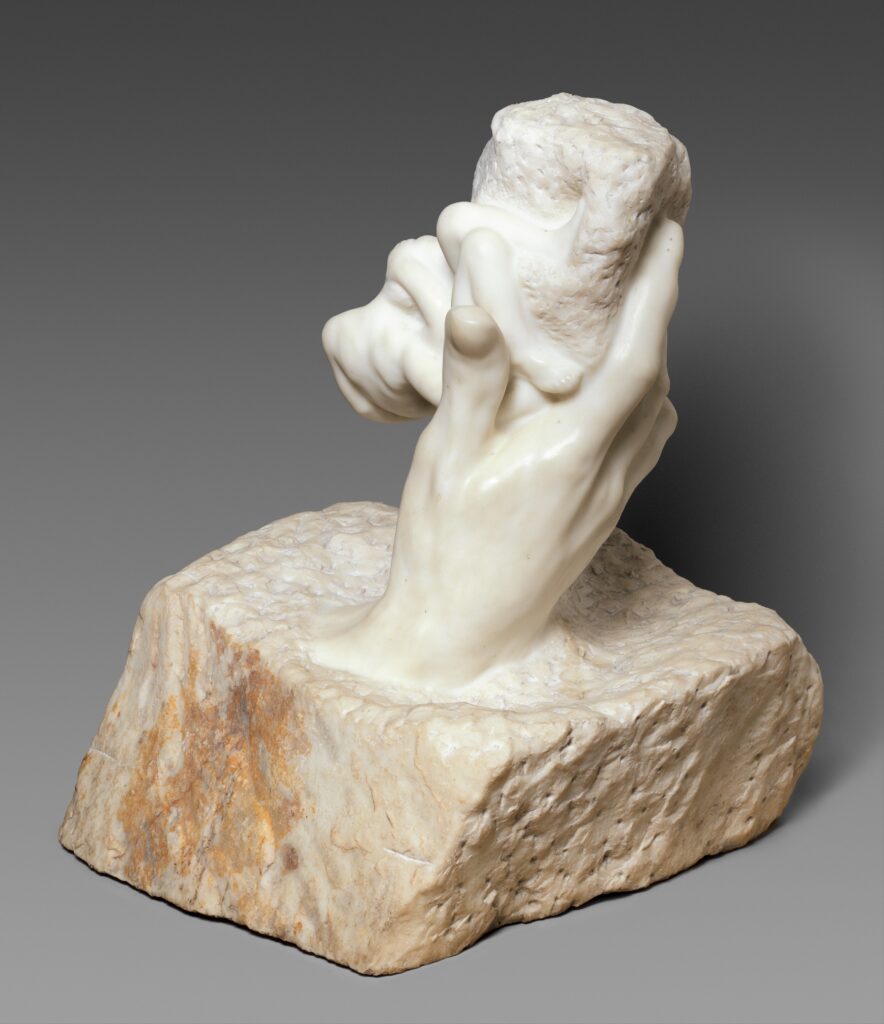 A marble sculpture depicts a hand emerging from a textured block of stone, grasping an indistinct, rough shape. The piece conveys a sense of creation.