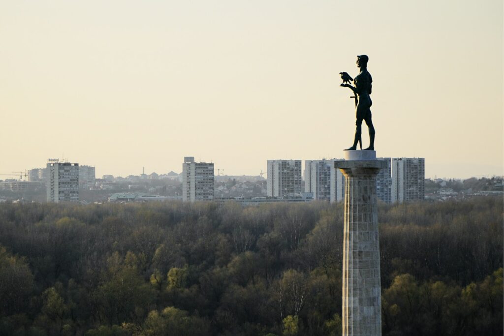 A statue of a man on a tall pedestal overlooks a forest with distant city buildings under a pale sky, conveying a sense of serenity and grandeur.