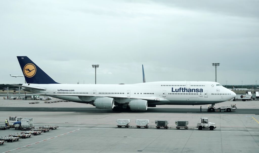 A Boeing 747-8 airplane on the tarmac under a cloudy sky, with a baggage cart and airport vehicles nearby. Calm, industrial setting at an airport.