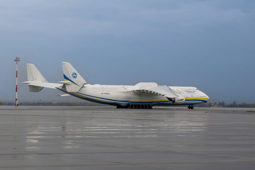 A massive Antonov cargo plane sits on a wet airport tarmac under a cloudy sky. The aircraft's large size dominates the scene, creating an imposing presence.
