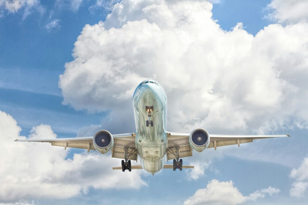 Airplane in flight viewed from below against a blue sky with scattered white clouds. The aircraft's landing gear is down, suggesting descent.