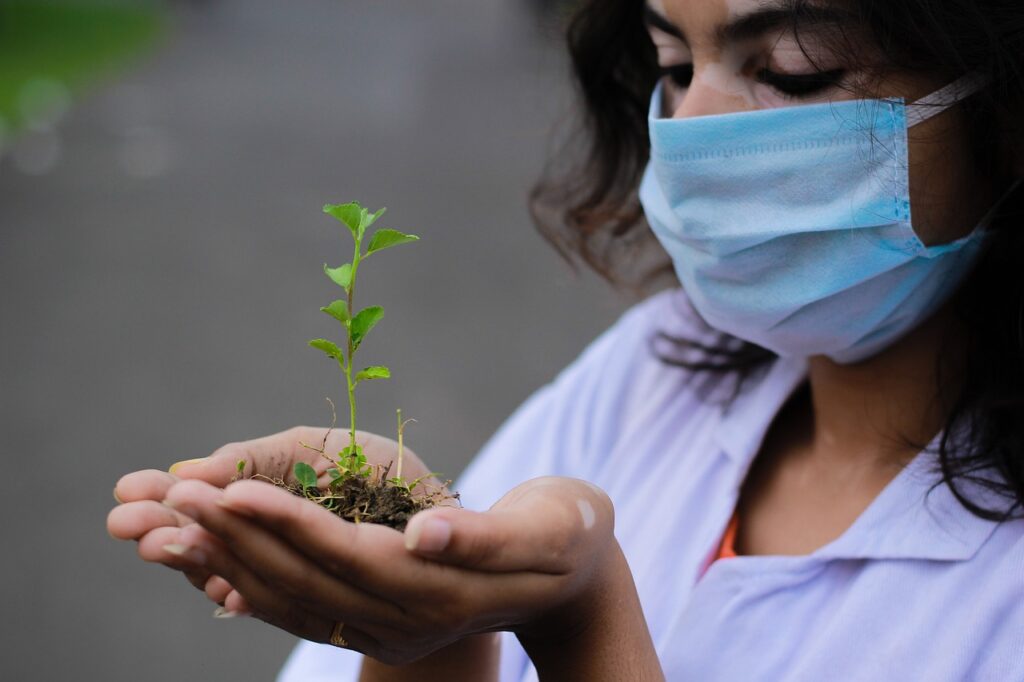 A woman in outdoor gear gently holds a young seedling in her hands, surrounded by lush greenery and dappled sunlight.