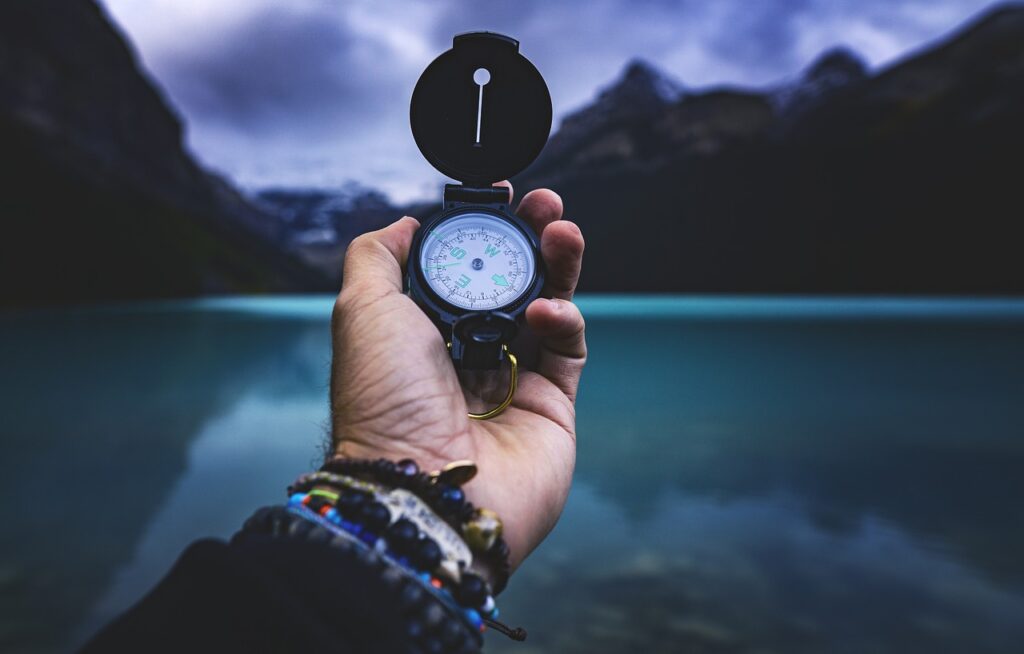 A person stands on a rocky cliff holding a compass, gazing toward distant mountains and a shimmering sea under a wide sky.