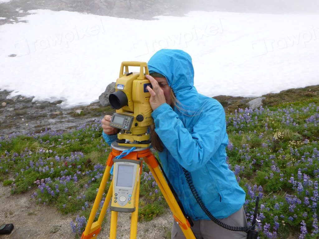 A field researcher kneels beside excavation tools and a laptop, analyzing data in a rugged outdoor environment surrounded by rocks and brush.